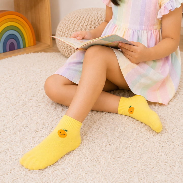 A child sitting on a white textured rug wearing a pastel rainbow-striped dress and bright yellow socks with a small orange pumpkin graphic. The child is holding an open book in a cozy room setting with a wooden shelf and rainbow decoration in the background.