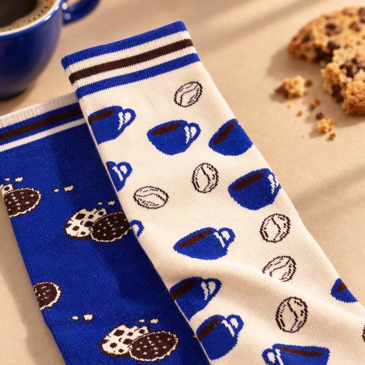 Two novelty crew socks displayed diagonally on a beige surface with coffee-themed props. One sock is royal blue with brown and white cookie patterns, and the other is off-white with blue coffee cup and coffee bean motifs.