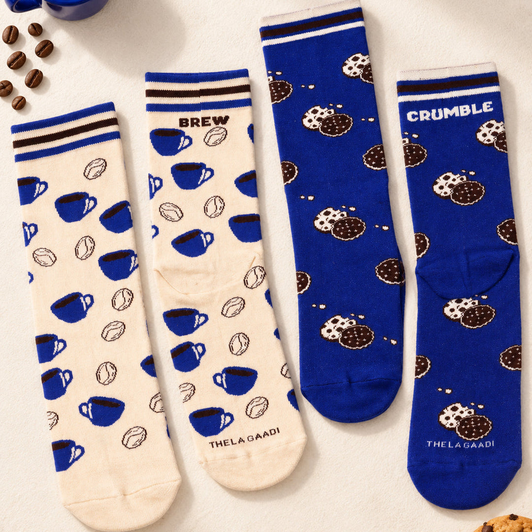 Four crew socks laid flat on a light cream background with coffee-themed props. Two socks are cream with blue coffee cup and bean prints, and two are royal blue with brown cookie and white cookie print.
